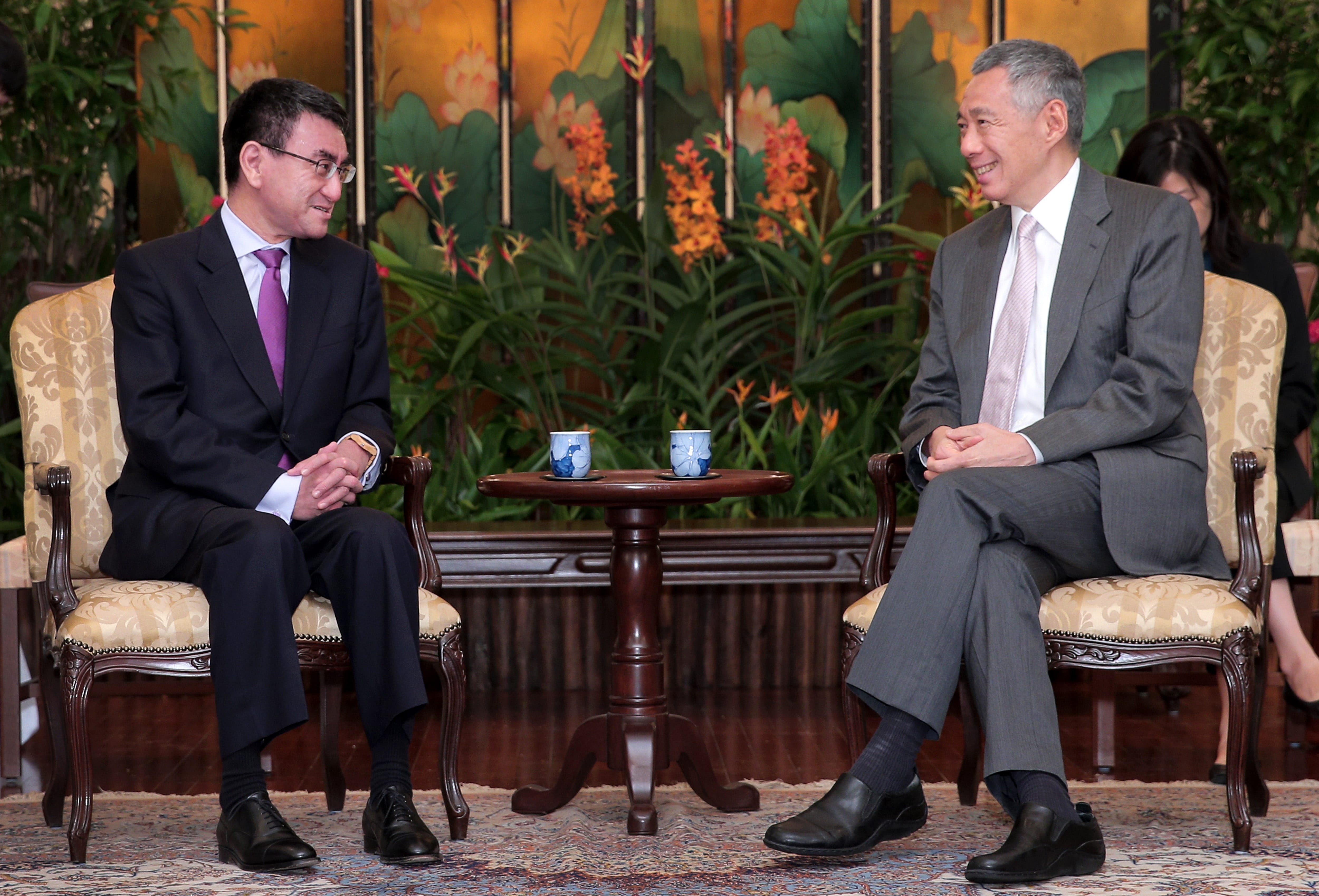 Two men in suits sit on chairs, facing each other across a small table with cups. Floral background.